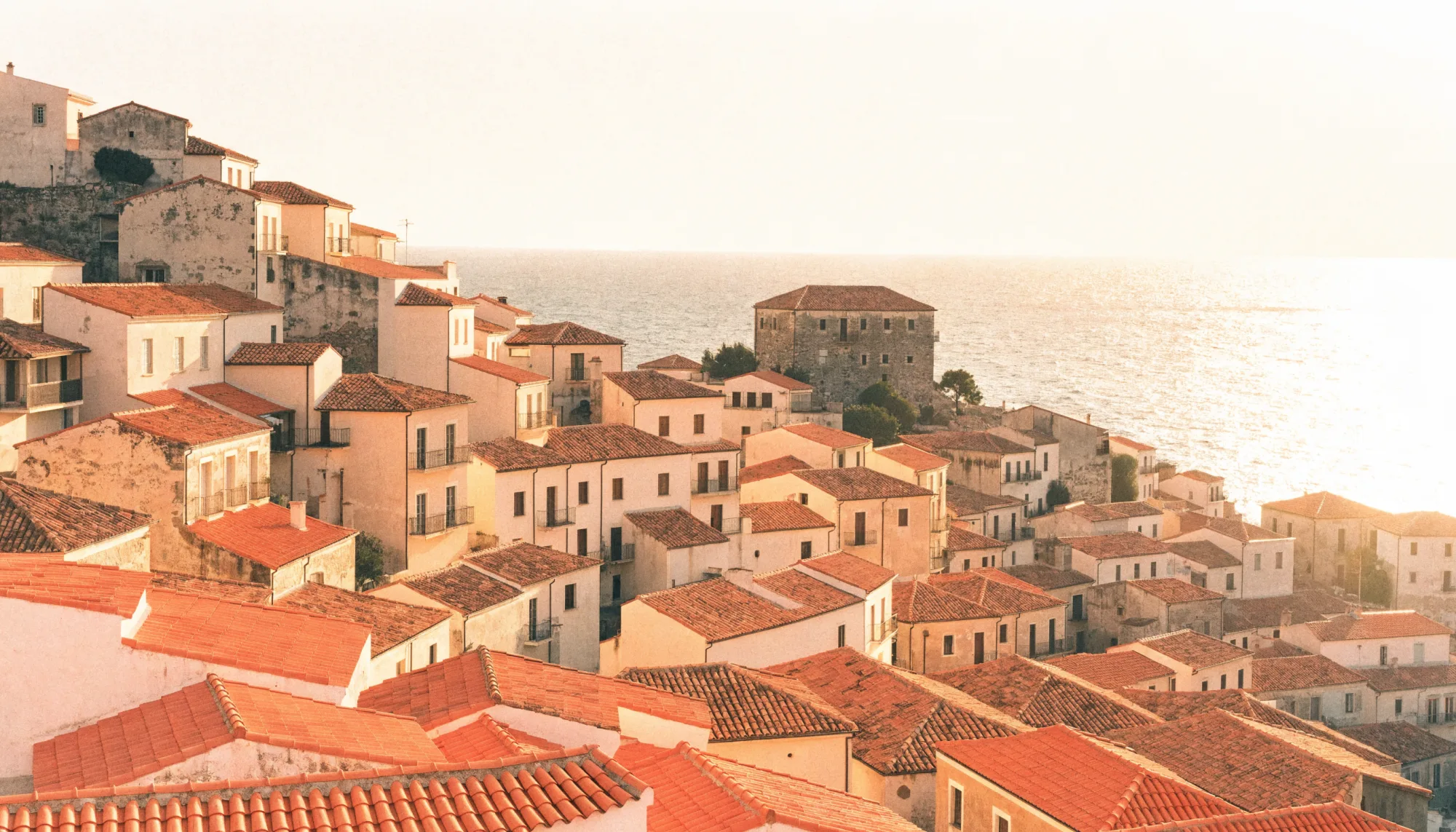 Terracotta rooftops of a hillside town stepping down to a calm sea at golden hour